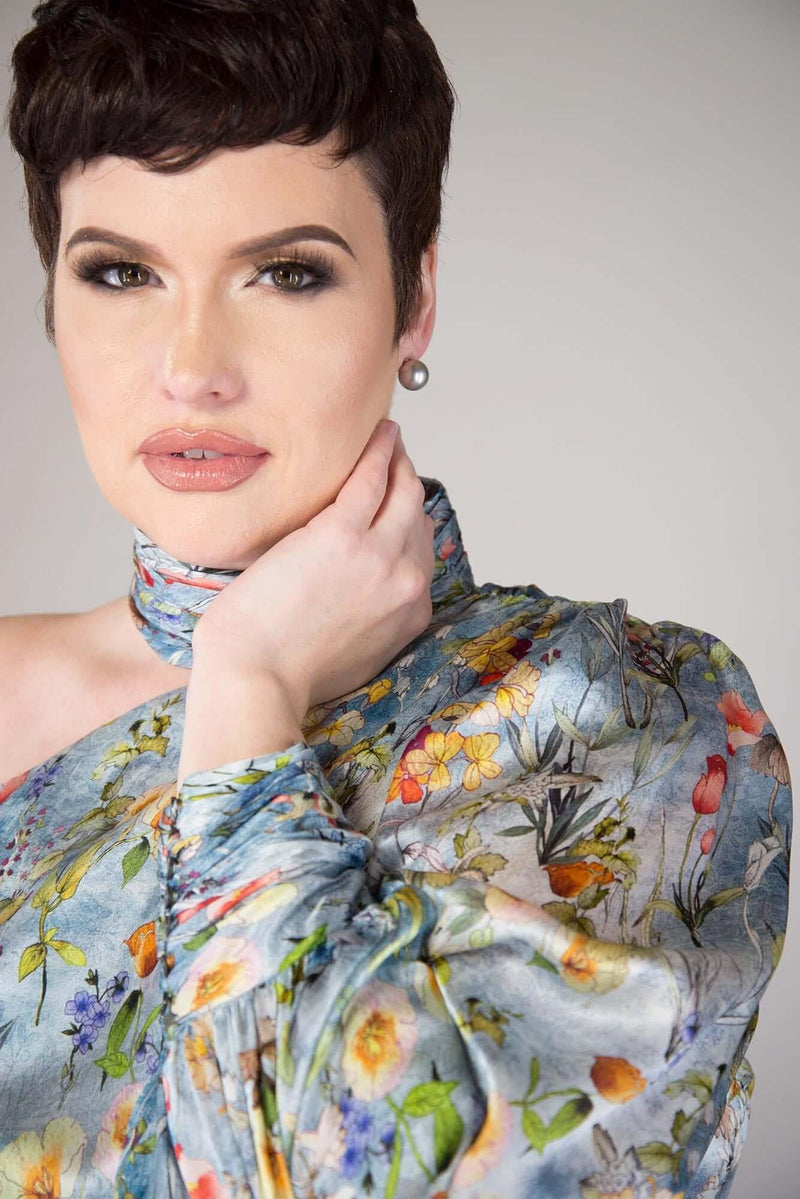 short haired model wears a Sky Blue, Botanical Flower Print one shouldered blouse with a Ruched Collar and High Cuff Volume Sleeve exposed shoulder blouse the details include: Four Button Collar, Seven Button High Cuff with elastic loops and full lined bodice.  Fabric is 100% Silk  garment is MADE IN NEW YORK CITY BY Mayes NYC